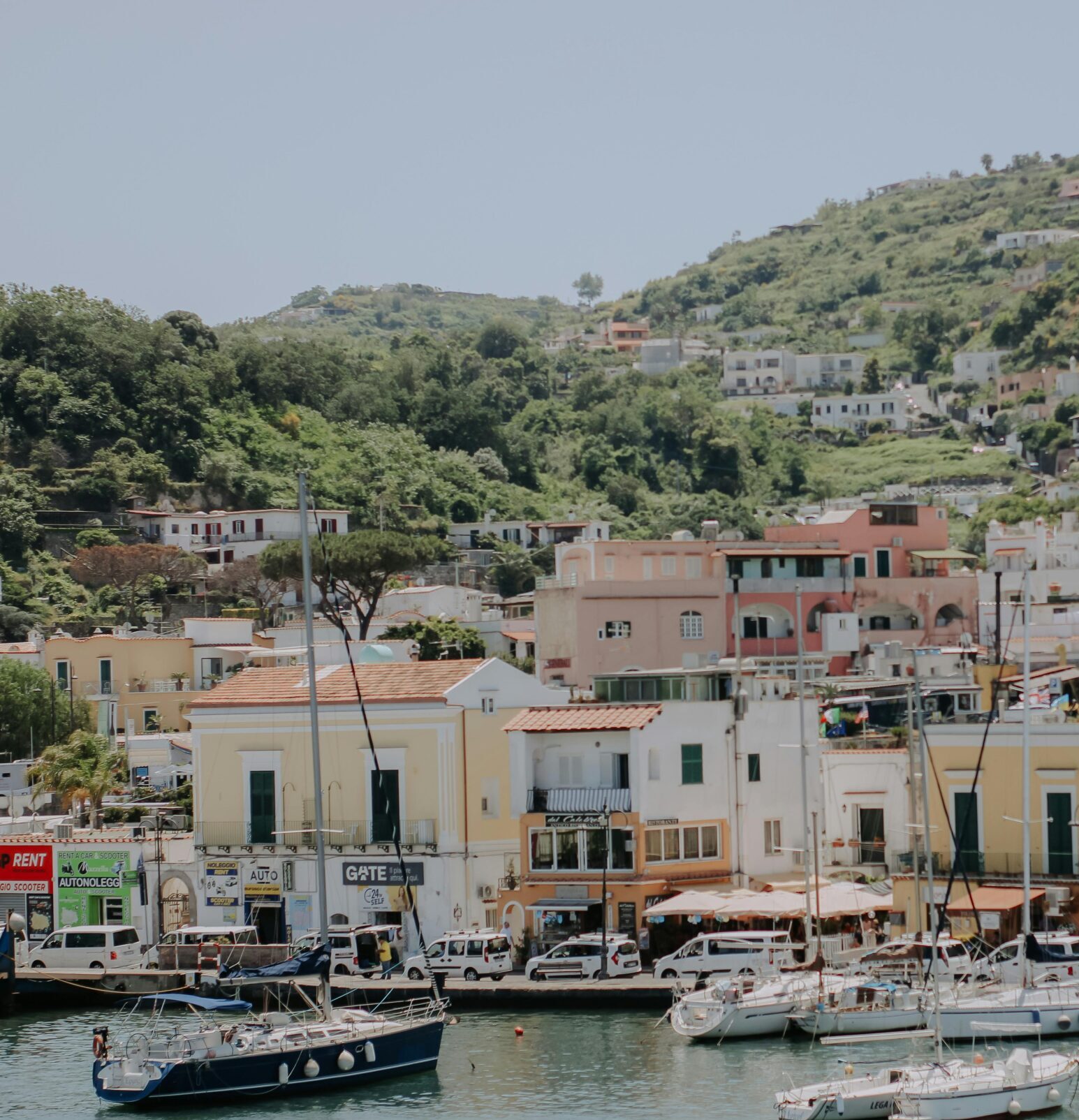 Scenic marina with sailboats and traditional hillside houses under clear blue skies.